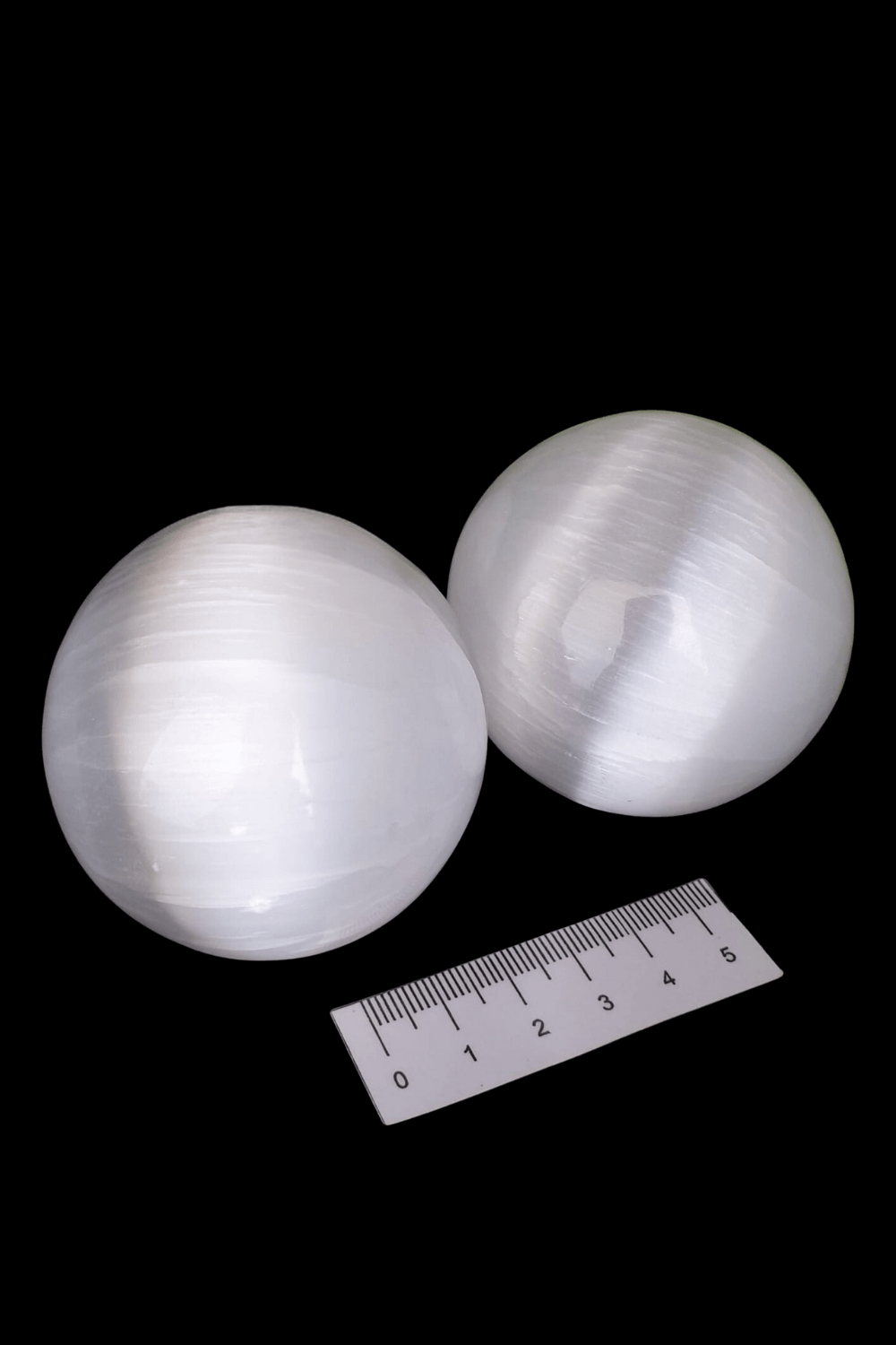 Two white selenite (Satin Spar) spheres with a black background, accompanied by a measuring ruler.